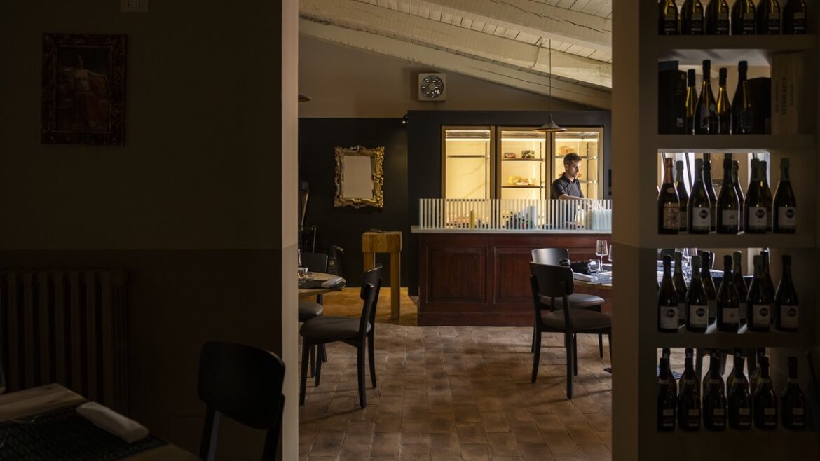 La sala di Tano Grano e Brace a Spello, con bancone illuminato, pavimento in cotto e cantina a vista