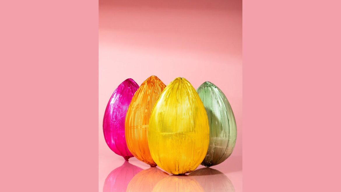 Quattro uova di Pasqua avvolte in carta stagnola colorata — rosa, arancione, giallo e verde — su sfondo rosa