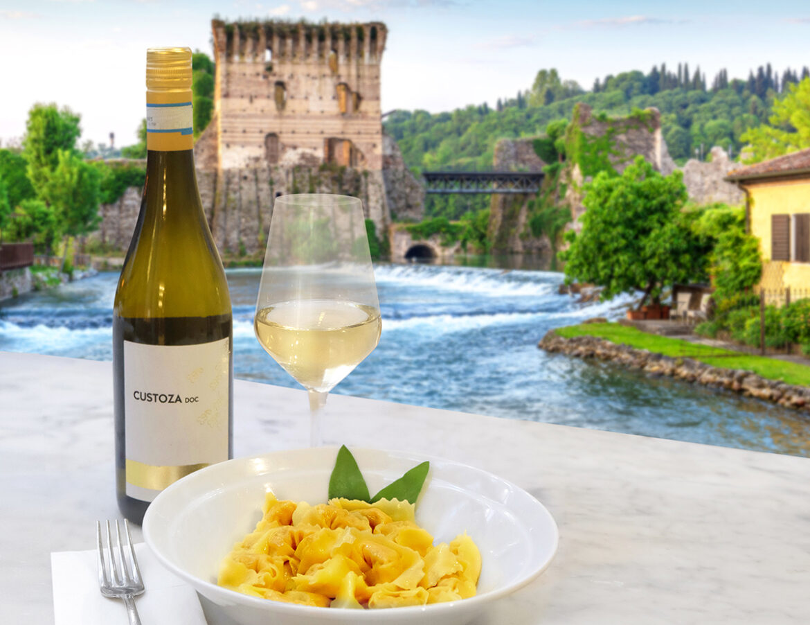 Un piatto di tortelli al Custoza Doc con una bottiglia di vino bianco e un calice su sfondo di Borghetto sul Mincio